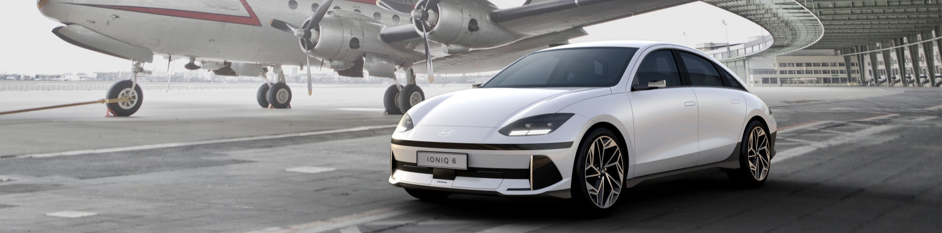 A white Hyundai IONIQ 6 standing on a road next to an airplane. The IONIQ 6 has a sleek aerodynamic silhouette and angular headlights.