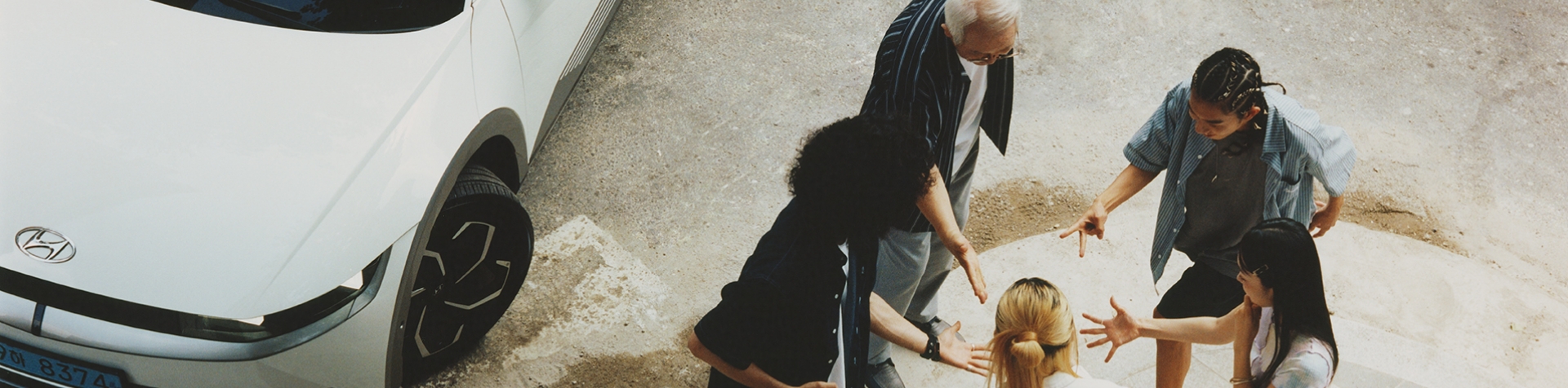 Birds’ eye view of an older man with 4 younger people standing in a circle and playing rock, paper, scissors with their hands in the middle. On the left-hand side, a white IONIQ 5 is parked. 