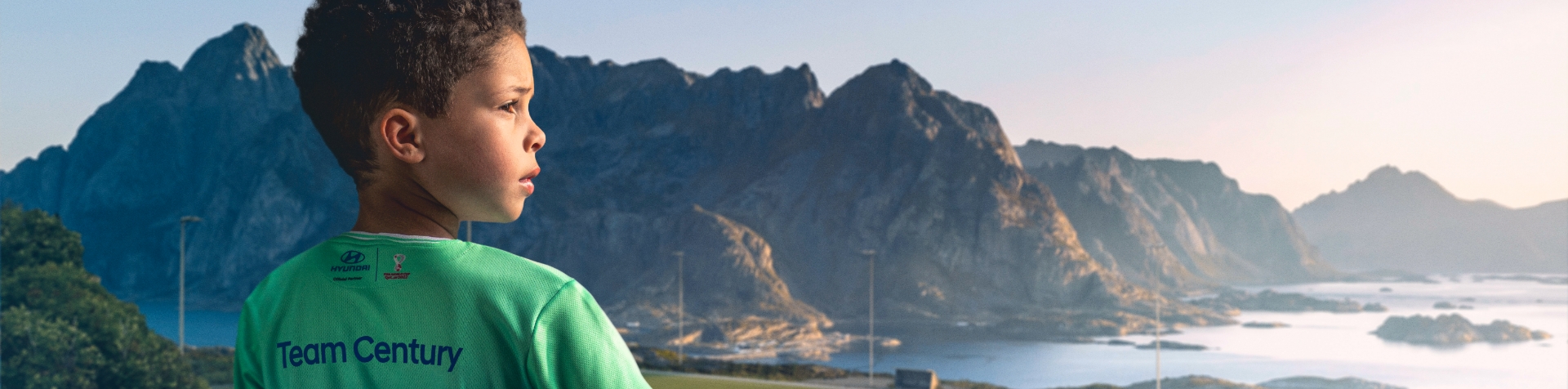 A young boy with dark hair wearing a Team Century jersey and looking out over a series of mountains and a lake from the top of a mountain.