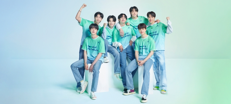 All 7 BTS members standing in a line facing forward. Each member has a green and gray Team Century shirt on with the Hyundai logo and Goal of the Century emblazoned in dark blue across the front.