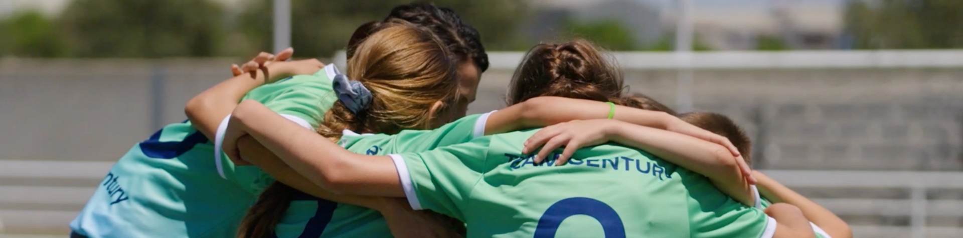 A group of children wearing green and blue Team Century jersey hug each other in a circle on the pitch.