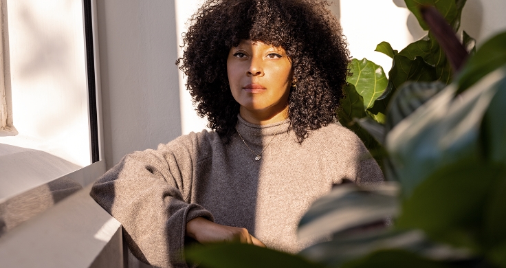 Nicky Woo sitting in a corner, with her elbow on a window ledge. She is lit by light from a window and surrounded by green plants. She is wearing a brown pull-over and a gold necklace.